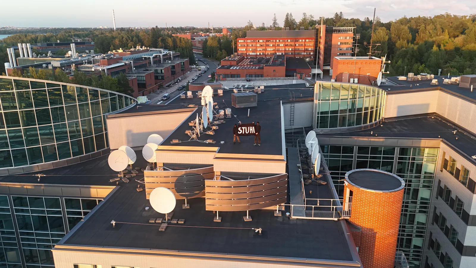 Two people in balaclavas hold a STUHI banner on the Aalto University rooftop at sunset